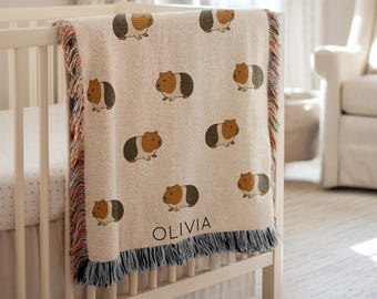 Puede incluir: Una manta de bebé tejida beige con un patrón repetido de ilustraciones de cobayas en marrón, negro y blanco. La manta tiene un borde de flecos multicolor y el nombre "OLIVIA" está impreso.