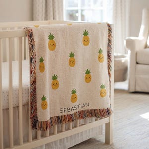 May include: A cream-colored baby blanket with a repeating pattern of smiling yellow pineapples with green tops. The blanket has a fringed edge with multiple colors and the name "SEBASTIAN" is printed in black.