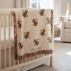 May include: A beige baby blanket with brown monkey illustrations and a fringed edge. The blanket is draped over a white crib. The name "OLIVIA" is printed on the blanket.