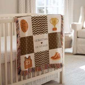 May include: A personalized baby blanket with a basketball theme. The brown and cream blanket features a basketball, trophy, jersey, and sneakers. The name "Sebastian" and the date "06-07-2025" are also displayed. The blanket is draped over a white crib.