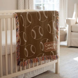 May include: A brown baby blanket featuring a baseball design with white detailing. The blanket, personalised with "CARTER JAMES", is draped over a white cot and has a fringed edge with a multi-coloured trim.