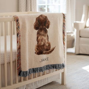 Puede incluir: Una manta de bebé beige tejida, con una ilustración de un perro marrón, sobre una cuna. La manta tiene un borde con flecos con hilos coloridos y el nombre "JAKE" impreso debajo.