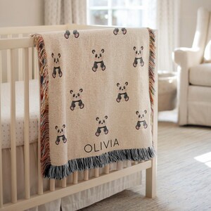 Peut inclure: Une couverture beige pour bébé ornée d'un motif répété de pandas noirs et gris. La couverture, avec une bordure à franges, est drapée sur un lit bébé blanc. Le nom "OLIVIA" est imprimé sur la couverture.