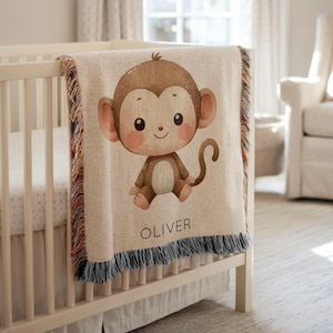May include: A beige baby blanket with a cartoon monkey illustration. The monkey is brown with large ears and a smiling face. The blanket has a fringe and the name "OLIVER" printed on it. The blanket is draped over a white crib.