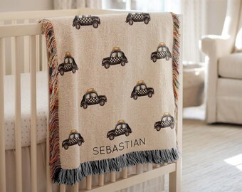 May include: A beige baby blanket with a repeating pattern of black and yellow taxi cabs. The blanket is draped over a white crib and has a fringe border. The name "SEBASTIAN" is printed on the blanket.