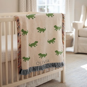 May include: A beige baby blanket with a repeating pattern of green lizards. The blanket is draped over a white crib and has a fringe border with multiple colors. The name "OLIVIA" is printed on the blanket.