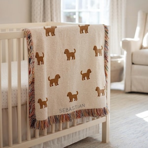 May include: A cream-coloured baby blanket featuring brown dog illustrations and the name "SEBASTIAN". The blanket has a fringed edge with multiple colours and is draped over a white cot.