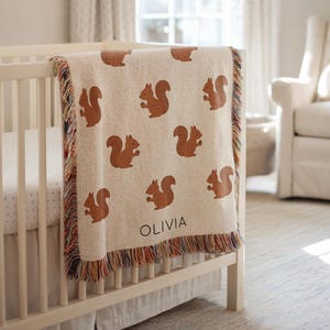 Pode incluir: Um cobertor de bebê bege com um padrão repetido de esquilos marrons. O cobertor está sobre um berço branco e tem uma borda com franjas multicoloridas. O nome "OLIVIA" é impresso no cobertor.
