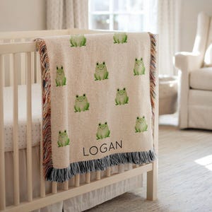 May include: A beige baby blanket with a repeating pattern of green frogs. The blanket is draped over a white crib. The name "LOGAN" is printed in black on the blanket. The blanket has a fringed edge with a mix of colors.