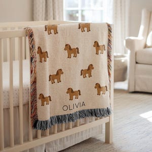 Op de afbeelding: Een beige babydeken met een herhaald patroon van bruine paarden. De deken heeft een franjerand en de naam "OLIVIA" is in het zwart gedrukt. De deken is gedrapeerd over een witte wieg in een kinderkamer.