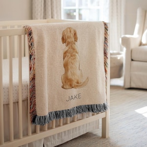 May include: A beige woven baby blanket with a watercolor illustration of a golden retriever puppy. The blanket has a fringed edge with red, blue, and orange threads. The name "JAKE" is printed in black letters on the blanket.