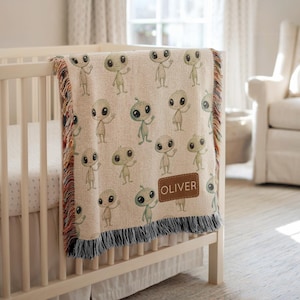 May include: A beige woven baby blanket featuring a repeating pattern of friendly green aliens. The blanket is draped over a white crib and has a brown patch with the name "OLIVER". The blanket has a fringed edge with multiple colors.