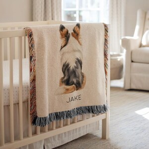 Puede incluir: Una manta tejida beige sobre una cuna, con una ilustración de acuarela de un perro desde atrás. El perro es blanco, marrón y negro, con el nombre "JAKE" impreso debajo. La manta tiene un borde con flecos.