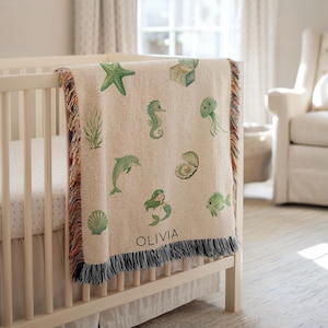 Op de afbeelding: Een beige babydeken met een zeedierenontwerp, waaronder een zeester, zeepaardje en zeemeermin. De deken heeft een franjebies en de naam "OLIVIA" erop gedrukt. Het ligt over een witte wieg.