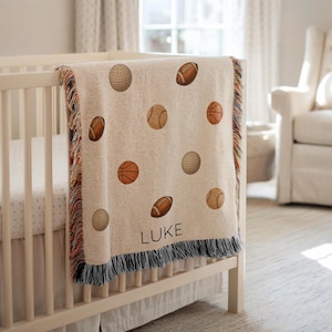 Puede incluir: Una manta de bebé beige con estampados de balones deportivos, incluyendo béisbol, fútbol americano y baloncesto. La manta tiene un borde con flecos y el nombre "LUKE" impreso. Está sobre una cuna blanca.