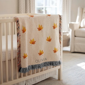 May include: A beige baby blanket with a repeating pattern of orange and yellow maple leaves. The blanket has a fringe border with a mix of colors and the name "OLIVIA" is printed on the blanket. The blanket is draped over a white crib.