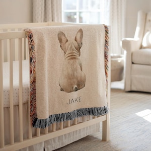 Könnte beinhalten: Eine beige Decke mit einem Aquarellbild des Hinterteils einer Französischen Bulldogge. Die Decke hat einen mehrfarbigen Fransenrand und den Namen "JAKE" in Schwarz aufgedruckt. Die Decke ist über ein weißes Kinderbett drapiert.