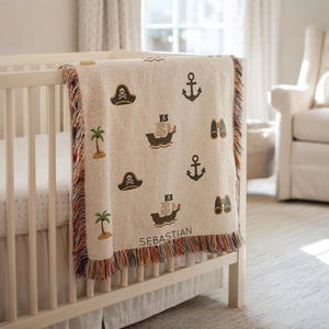 May include: A beige baby blanket with a pirate theme, draped over a white cot. The blanket features images of pirate ships, anchors, eye patches, palm trees, and binoculars. The name "SEBASTIAN" is printed on the blanket.