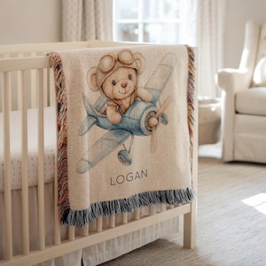 Puede incluir: Una manta de bebé beige con una ilustración de un oso de peluche con gorro y gafas de aviador, pilotando un avión azul. La manta tiene un borde con flecos y el nombre "LOGAN" está impreso.