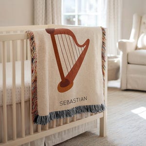 May include: A beige baby blanket with a brown harp design and the name "SEBASTIAN" in black lettering. The blanket has a fringed edge with multiple colors and is draped over a white crib.
