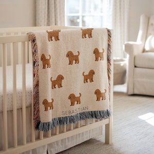 May include: A beige woven baby blanket with a repeating pattern of brown dog illustrations. The blanket is draped over a white crib and has a fringe border. The name "SEBASTIAN" is woven into the blanket.