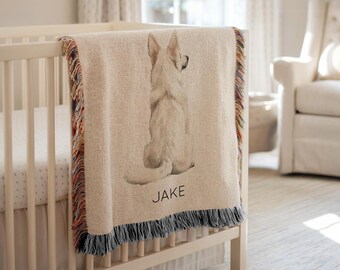 May include: A beige throw blanket featuring a watercolour image of a dog draped over a cot. The blanket has a fringed edge with multiple colours. The name "JAKE" is printed in black at the bottom of the blanket.