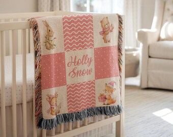 May include: A pink and beige baby blanket with a patchwork design featuring woodland animals and the words "Holly Snow." The blanket has a fringed edge and is draped over a white cot. The blanket has a dotted and wavy pattern.