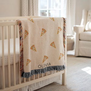 May include: A beige baby blanket with a repeating pattern of pizza slices. The blanket is draped over a white crib. The name "OLIVIA" is printed in black on the blanket. The blanket has a fringed edge.