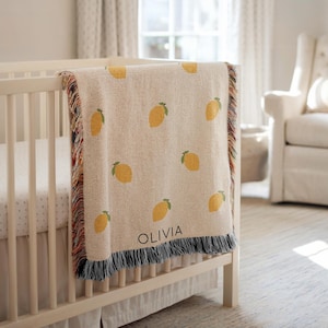 May include: A cream-coloured baby blanket featuring a pattern of yellow lemons and green leaves. The blanket, with a fringed border, is draped over a white cot. The name "OLIVIA" is printed in black on the blanket.