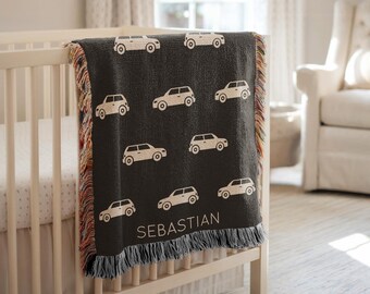 May include: A dark gray baby blanket with a repeating pattern of white cars. The blanket is draped over a white crib and has a fringe border with a personalized name, "SEBASTIAN," in white.