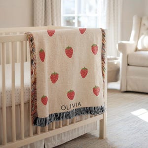 May include: A beige baby blanket with a repeating pattern of red strawberries and green leaves. The blanket is draped over a white crib and has a fringe border. The name "OLIVIA" is printed on the blanket.