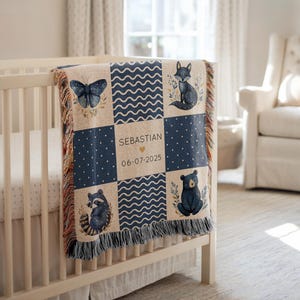 May include: A personalised baby blanket with a patchwork design featuring a butterfly, fox, raccoon, and bear. The blanket is navy blue and beige with the name "SEBASTIAN" and the date "06-07-2025" printed on it. The blanket is draped over a white cot.