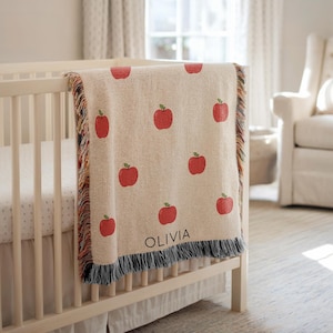 May include: A cream-colored baby blanket with a repeating pattern of red apples and green leaves. The blanket is draped over a white crib and has a fringe border. The name "OLIVIA" is printed in black on the blanket.