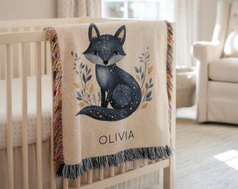 May include: A beige baby blanket with a navy blue fox illustration, surrounded by leaves and stars. The blanket has a fringed edge and the name "OLIVIA" printed in black. The blanket is draped over a white crib.