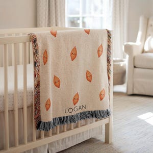 May include: A beige baby blanket featuring a pattern of orange sweet potatoes with smiling faces. The blanket has a fringed border and the name "LOGAN" is printed in black. The blanket is draped over a white cot.