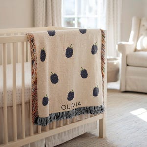 May include: A beige baby blanket with a pattern of dark blue blackberries and green leaves. The blanket is draped over a white crib and has the name "OLIVIA" printed on it. The blanket has a fringed edge.