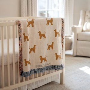 May include: A cream-coloured baby blanket featuring a repeating pattern of golden-brown dog silhouettes. The blanket is draped over a white cot and has a fringed border with multiple colours. The name "SEBASTIAN" is printed on the blanket.