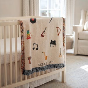 Può includere: Coperta beige tessuta con grafiche di strumenti musicali colorati, tra cui una chitarra, un pianoforte e un sassofono. La coperta ha un bordo a frange e il nome "SEBASTIAN" è stampato sopra.