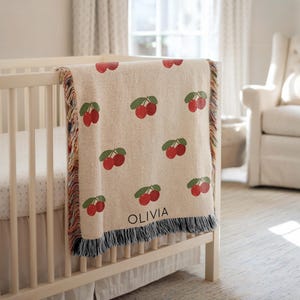 Könnte beinhalten: Eine beige Babydecke mit einem sich wiederholenden Muster aus roten Kirschen und grünen Blättern. Die Decke ist über ein weißes Kinderbett drapiert und hat einen Fransenrand. Der Name "OLIVIA" ist in schwarzen Buchstaben auf die Decke gedruckt.
