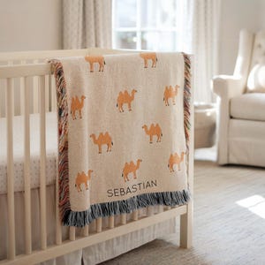 May include: A beige woven baby blanket with a repeating pattern of orange camels. The blanket is draped over a white crib and has a fringe border with the name "SEBASTIAN" printed on it.