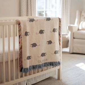 以下が含まれることがあります： ベージュ色の織りベビーブランケット。グレーのオポッサムの繰り返し模様。 ブランケットは、グレー、赤、オレンジ色の糸のフリンジボーダーが付いています。 「OLIVIA」という名前がグレーの文字でブランケットに印刷されています。 ブランケットは白いベビーベッドに掛けられています。