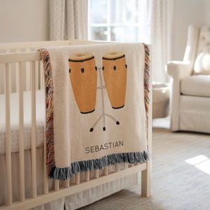 Op de afbeelding: Een beige babydeken met een kleurrijke franje en een zwart-witte franje. De deken heeft een afbeelding van twee bruine conga-drums op een standaard, met de naam "SEBASTIAN" eronder afgedrukt.