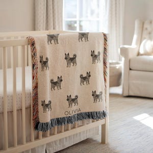 May include: A beige woven blanket with a repeating pattern of gray dog illustrations. The blanket is draped over a white crib and has a fringe border with multiple colors. The name "OLIVIA" is printed on the blanket.