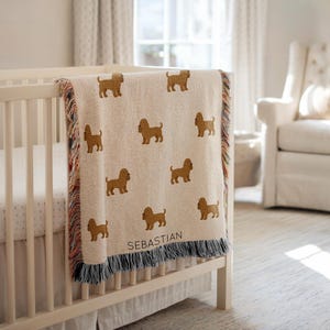 Può includere: Una coperta per bambini beige con un motivo ripetuto di sagome di cani marroni. La coperta è drappeggiata su una culla bianca e ha un bordo a frange multicolore. Il nome "SEBASTIAN" è stampato sulla coperta.