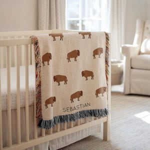 May include: A beige woven baby blanket with a repeating pattern of brown bison. The blanket is draped over a white crib and has a fringe border with multiple colors. The name "SEBASTIAN" is printed on the blanket.