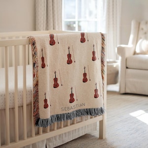 May include: A cream-colored baby blanket with a repeating pattern of red violins and bows. The blanket is draped over a white crib and has a fringed edge. The name "SEBASTIAN" is printed on the blanket.