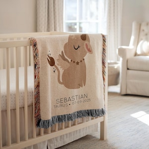 Könnte beinhalten: Beige Babydecke mit einer Cartoon-Stier-Illustration, Flügeln und einer Sternenkette. Die Decke hat einen Fransenrand und den Text "Sebastian, Taurus, 07-09-2025". Die Decke ist über ein weißes Kinderbett drapiert.