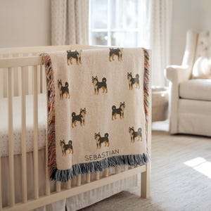 May include: A beige woven blanket with a repeating pattern of black and brown dogs. The blanket has a fringe border with red, orange, and blue threads. The name "Sebastian" is printed on the blanket.