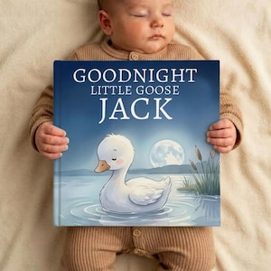 May include: A baby lies peacefully, holding a children's book. The book cover features a serene illustration of a white goose on a lake under a full moon. The title reads "GOODNIGHT LITTLE GOOSE JACK". The baby is wearing a brown onesie.