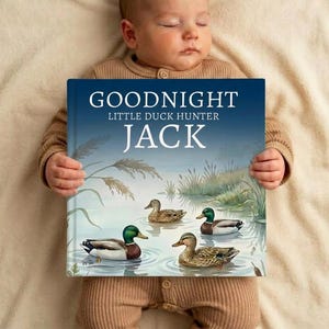 Op de afbeelding: Een baby in een bruine onesie houdt een kinderboek vast met de titel "Goodnight Little Duck Hunter Jack". De kaft van het boek bevat illustraties van eenden die in een vijver zwemmen. De baby slaapt op een crèmekleurige deken.